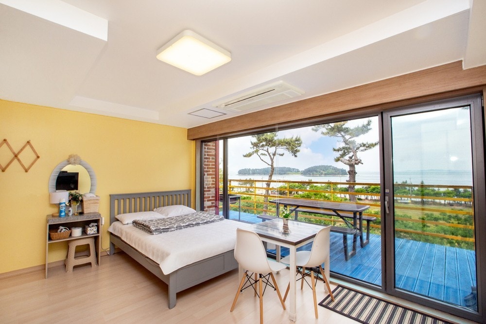 A brightly lit room features a spacious bed and a small seating area with a table and chairs. Large glass doors open to a deck area, offering views of the sea and surrounding trees, while a wall mirror adds a touch of elegance.