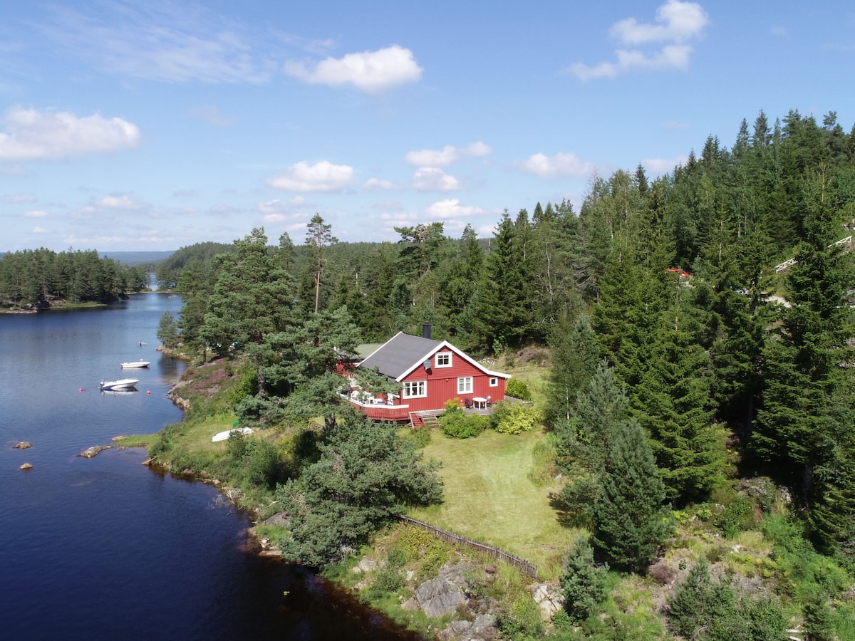 A detached red house is nestled among lush greenery by a serene lake, surrounded by trees. The natural landscape includes a grassy area leading to the water's edge, with several boats visible in the calm lake, enhancing the tranquil setting.