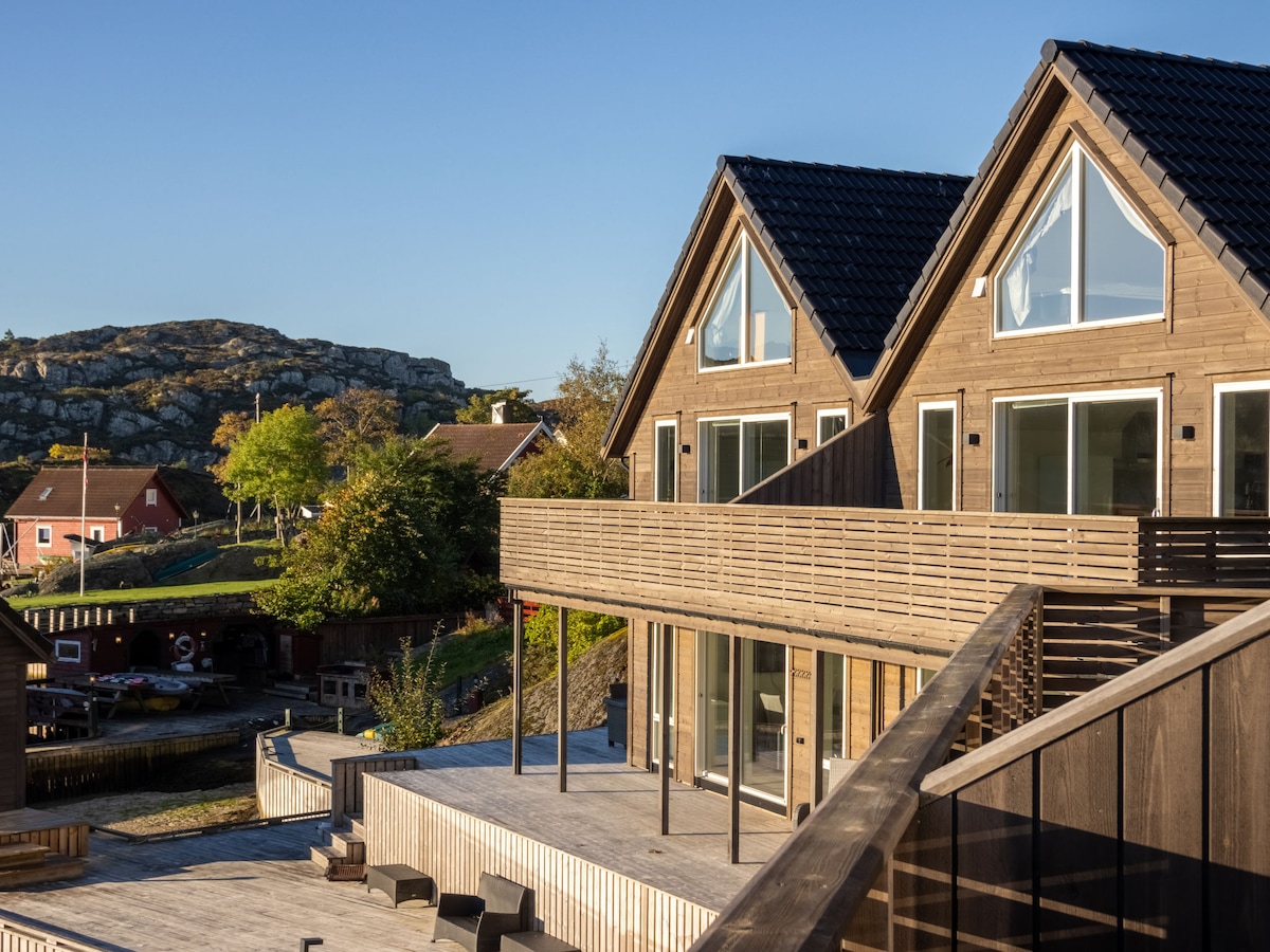 The exterior showcases a modern semi-detached house with large glass windows and a spacious wooden terrace. Surrounding greenery complements the structure, while a rocky hill rises in the background. The patio area features outdoor seating, enhancing the connection to the natural landscape.