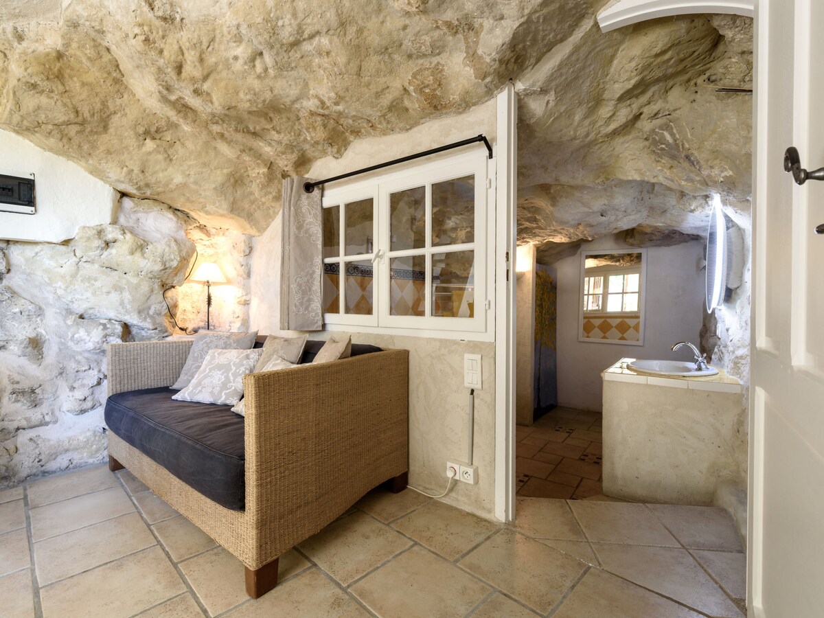 A cozy living area is set against a natural stone wall, featuring a comfortable sofa with decorative cushions. White-framed windows provide natural light, and a small sink area is visible in the background. The floor is tiled with light-colored ceramic tiles.