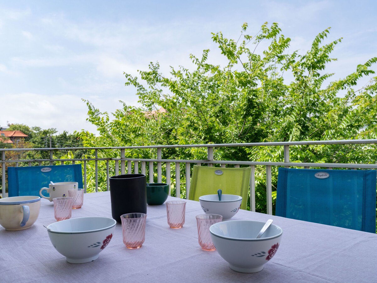 A dining table is set on a terrace, featuring a white tablecloth and various bowls and glasses. Colorful chairs in shades of blue, green, and pink surround the table. Lush greenery provides a natural backdrop, enhancing the outdoor dining experience.
