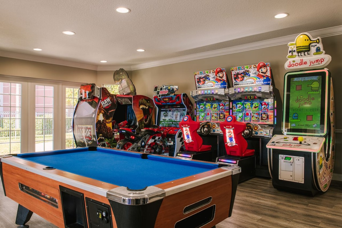 A game room is equipped with various arcade machines and a pool table. Brightly colored machines include racing games, a Doodle Jump arcade unit, and classic arcade designs, offering a fun and engaging atmosphere for guests of all ages.