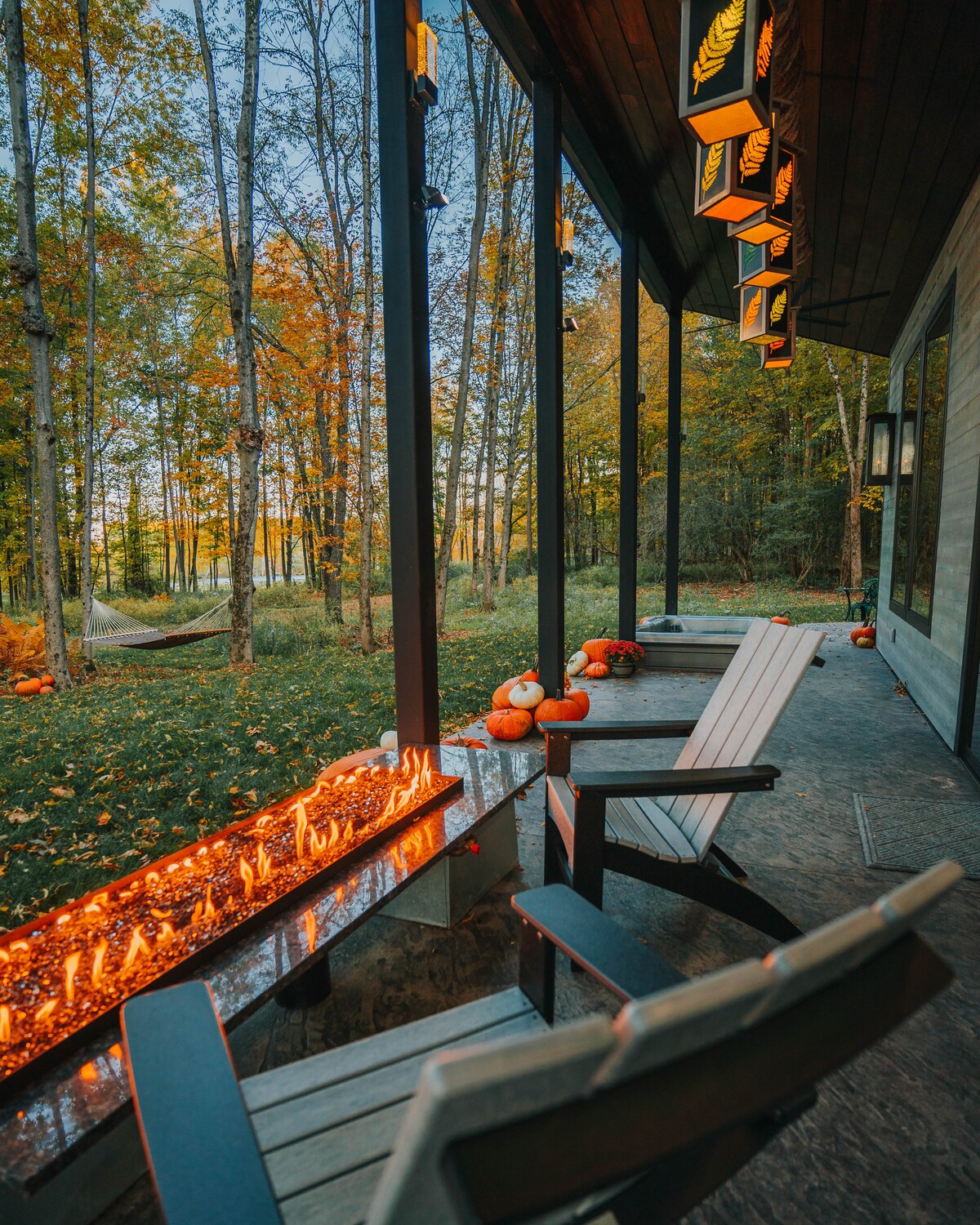 A serene outdoor space features two wooden benches positioned beside a fire table. Warm flames flicker along a stone edge, surrounded by seasonal decorations like pumpkins. Tall trees with autumn foliage create a peaceful backdrop, adding a touch of nature to the atmosphere.