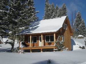 The Smith Cabin, constructed with natural wood, is framed by snow-covered trees. The front porch, equipped with a grill, is visible beneath a gently sloping roof. Large windows allow for natural light, while the surrounding snow creates a serene winter landscape.
