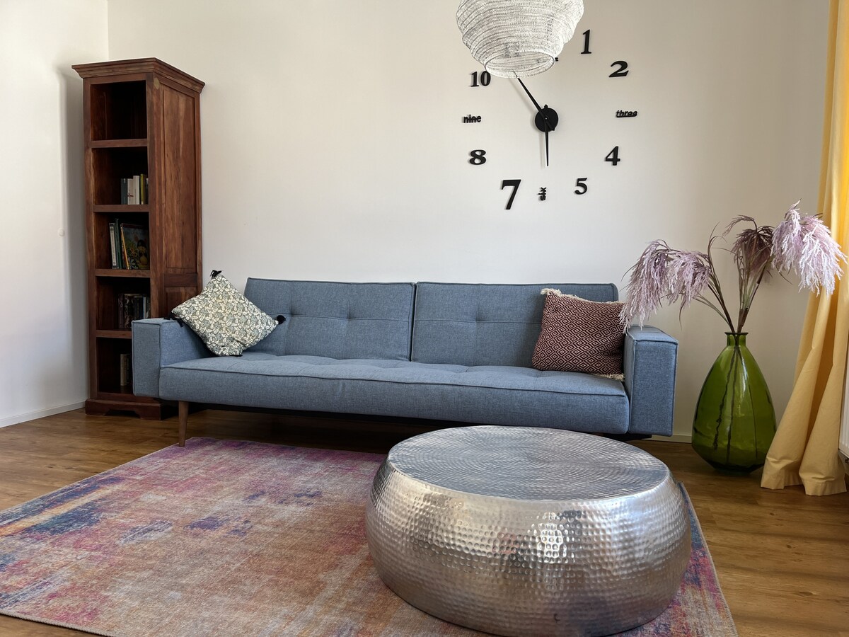 A modern living area features a comfortable gray sofa adorned with patterned cushions. A decorative round coffee table with a metallic finish sits on a colorful area rug. A tall bookshelf stands nearby, and a large wall clock serves as a focal point above the sofa.