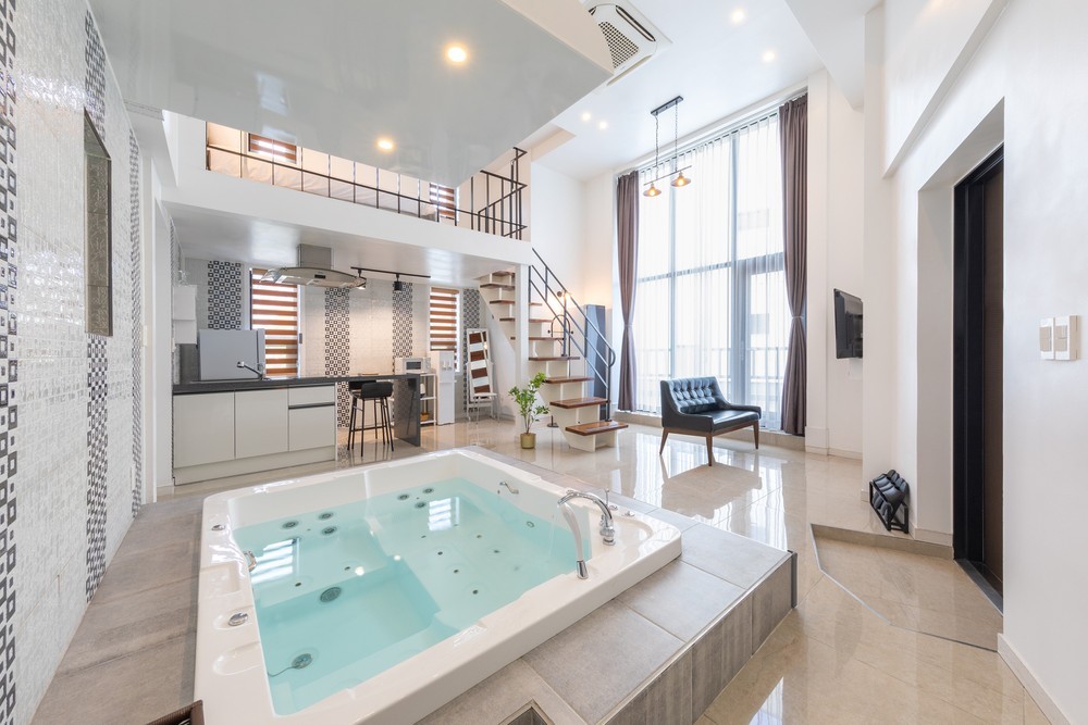 A spacious interior showcases a large whirlpool tub at the forefront, surrounded by polished tiles. The open layout reveals a staircase leading to an upper level, with a modern kitchen and seating area visible in the background. Large windows allow natural light to fill the room.