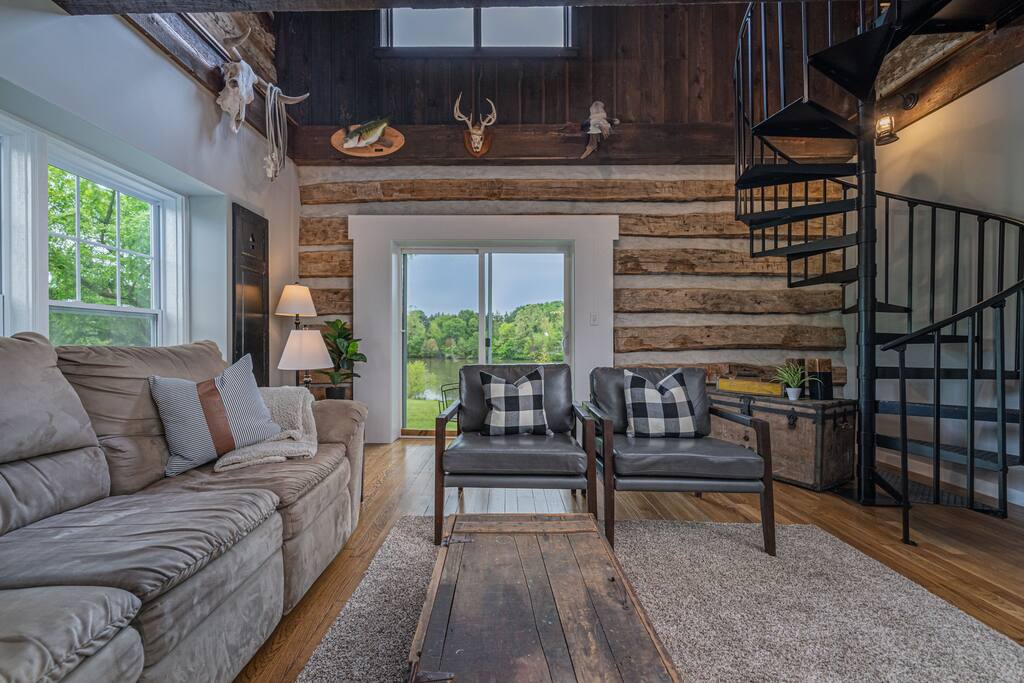 A cozy living area features a large, comfortable couch and two armchairs with plaid cushions. Natural light floods through large windows, offering a view of the lake. A spiral staircase leads to the loft, surrounded by rustic wood paneling and decorative mounted accents.