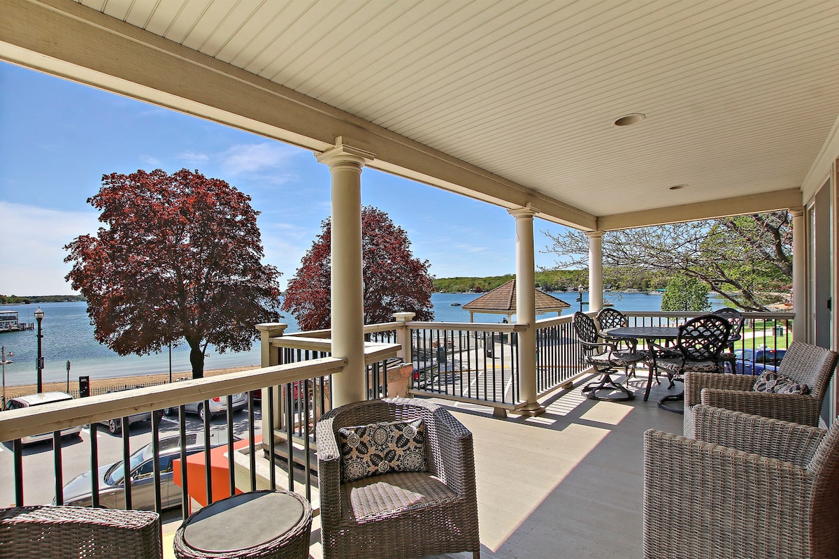 A spacious deck overlooks a serene lake, framed by lush trees. Several comfortable wicker chairs surround a glass-topped table, inviting relaxation. The railing provides a clear view of the water, while shadow and sunlight interplay on the surface, showcasing the surrounding natural beauty.