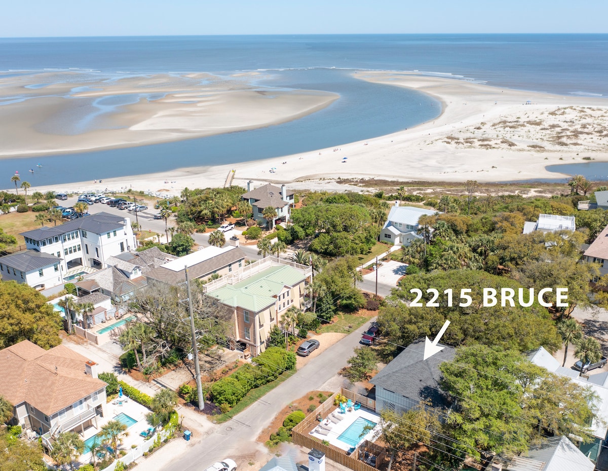 An aerial view of the neighborhood surrounding Three Oaks Cottage, showing the proximity to the beach and waterways. The beachfront is visible with soft sandy areas and gentle waves. Nearby trees and houses create a serene coastal atmosphere, with the location marked as 2215 Bruce.