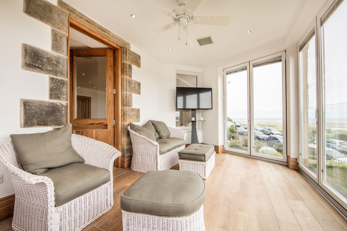 The sunroom features comfortable wicker seating with neutral cushions, complemented by a matching ottoman. Large glass bi-fold doors open to the Juliet balcony, offering expansive views of the sea and surrounding landscape. Natural light floods the space, highlighting the warm wood flooring.