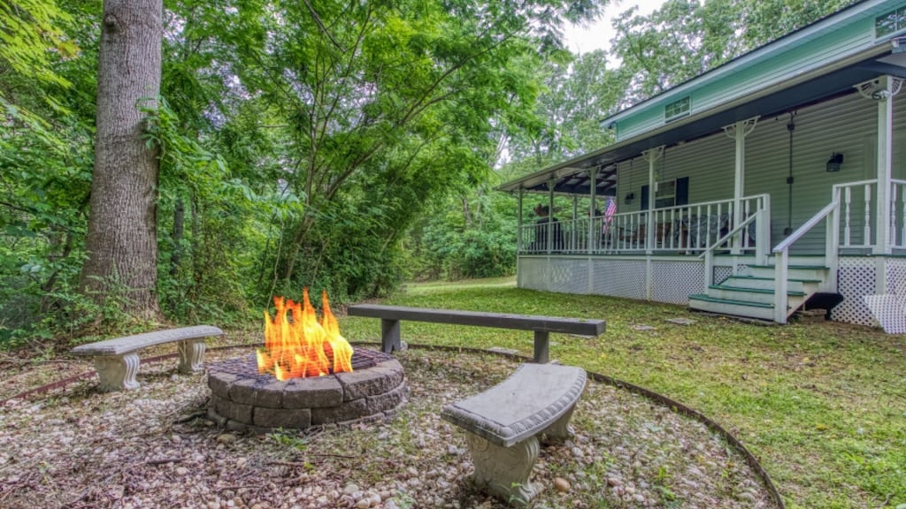 A firepit area surrounded by stone seating is set within a grassy space. Lush green trees provide a natural backdrop, while the spacious wraparound porch of The Farmhouse is visible in the background, inviting relaxation amidst the serene outdoor setting.