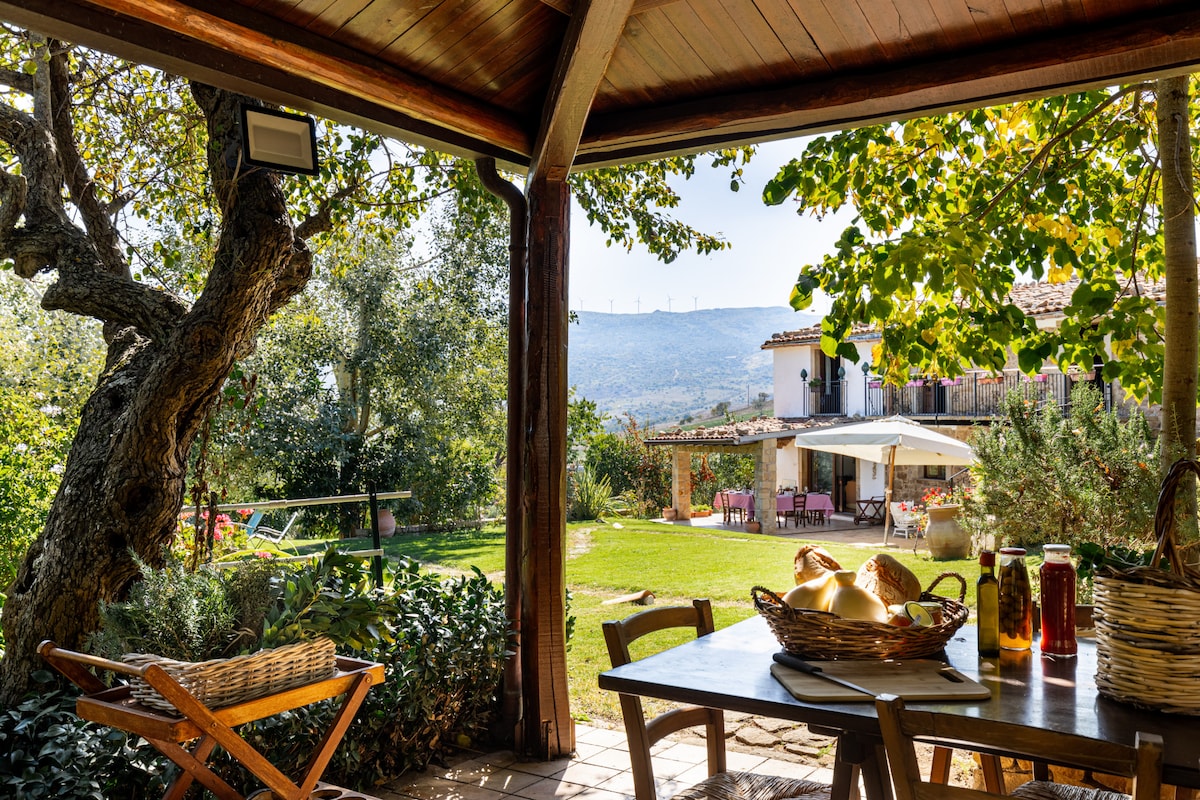 An outdoor dining area features a wooden table with woven baskets, surrounded by greenery. A sun umbrella provides shade, and a spacious lawn is visible in the background, highlighting the serene landscape with distant hills and a charming building.