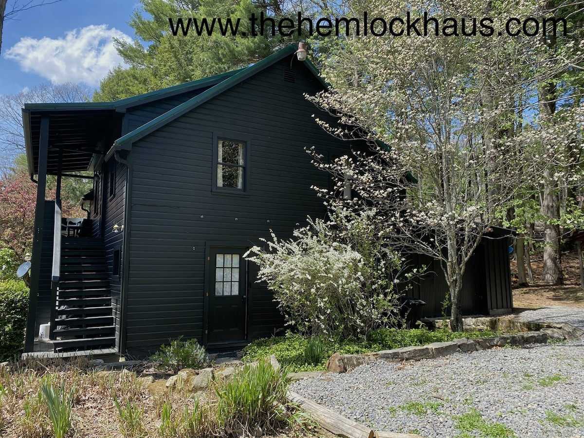 A black wooden cabin is shown surrounded by lush greenery and blooming trees. The structure features multiple levels, with an exterior staircase leading to an upper entrance. A gravel area is visible in the foreground, complementing the natural setting.