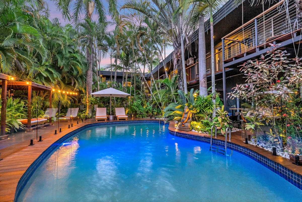 A private pool oasis is surrounded by lush tropical greenery, featuring oversized sunlounges and a shaded cabana. Soft lighting along the boardwalk illuminates the tranquil atmosphere, with the water reflecting the colors of the evening sky.