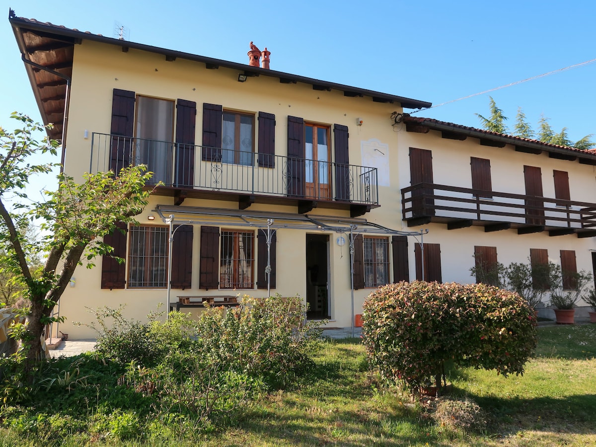 The semi-detached house features a light-colored exterior with dark shutters and a balcony. A covered patio area is visible, accompanied by garden furniture. Surrounding greenery includes well-kept shrubs and trees, creating a welcoming outdoor space.