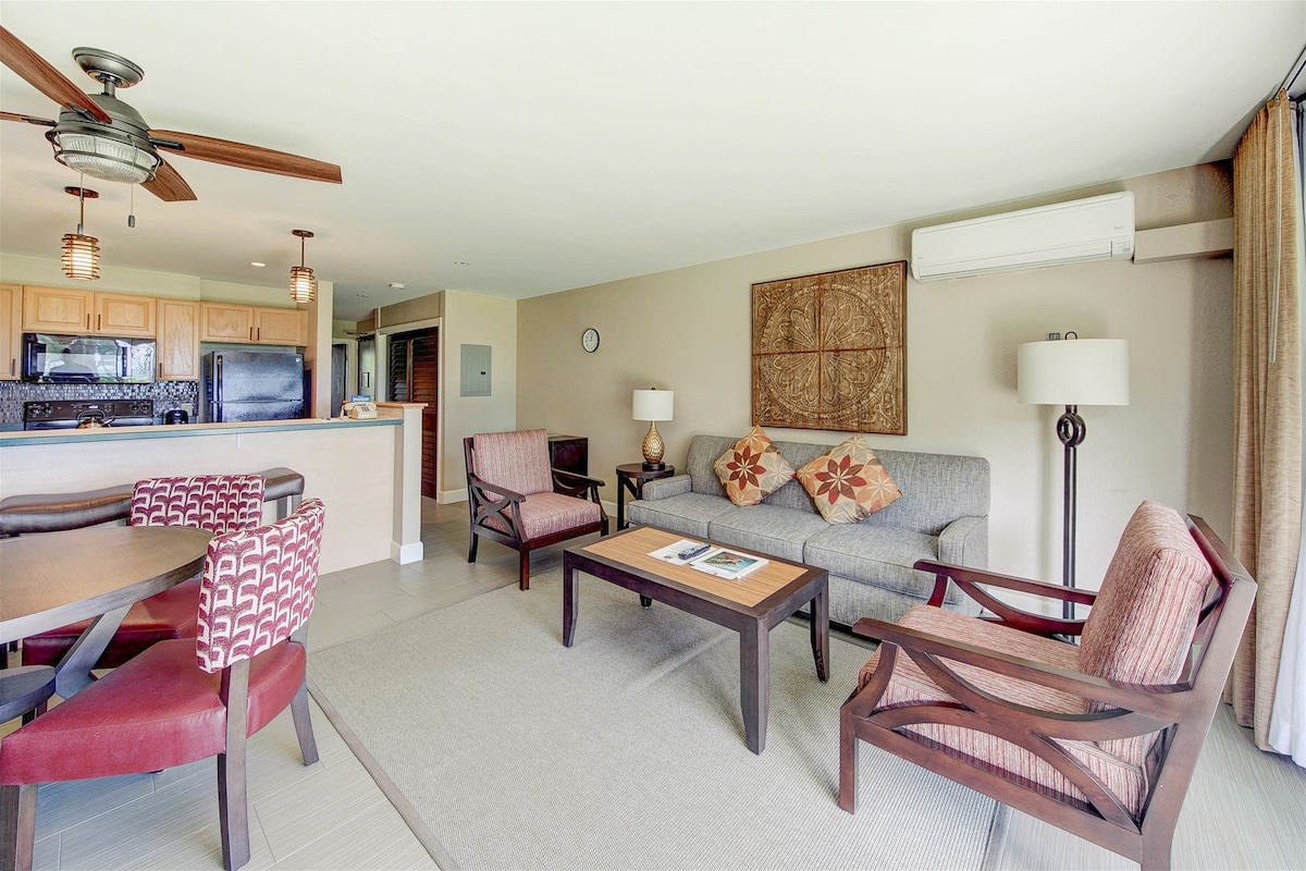 A comfortable living area is highlighted by a cozy sofa with decorative cushions. Two armchairs and a round dining table provide ample seating. A well-equipped kitchen is accessible in the background, and sunlight filters through the large windows, enhancing the open and airy atmosphere.