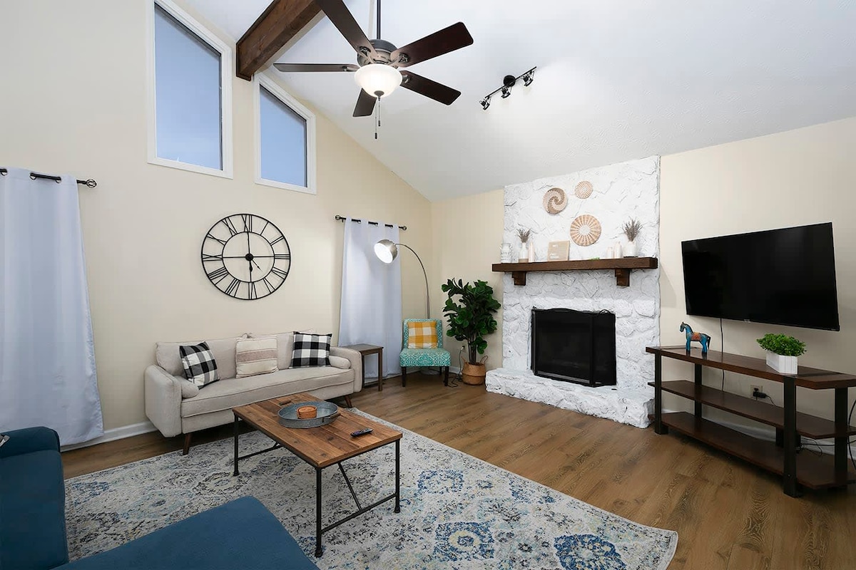 A cozy living area features a light-colored sofa paired with a patterned area rug. A stone fireplace serves as a focal point, complemented by a wooden mantel adorned with decorative items. Large windows allow natural light, and a television is mounted nearby.