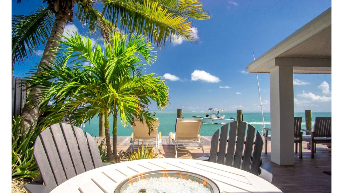 A private outdoor space showcases a circular fire pit surrounded by four comfortable chairs. In the background, palm trees frame a view of the ocean, with a boat seen cruising in the distance under a clear blue sky.
