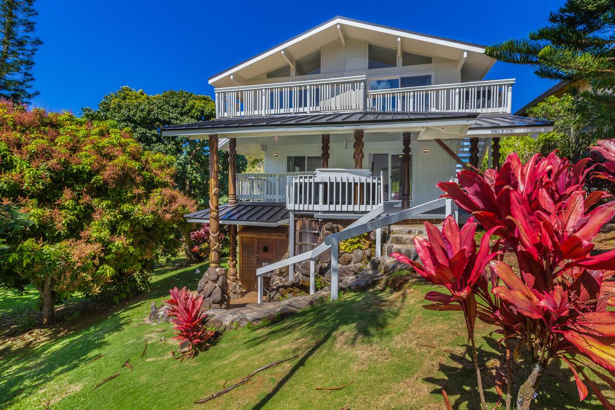 The exterior of a modern two-story home is surrounded by lush greenery and vibrant tropical plants. A wide staircase leads to a covered lanai at the second level, which features railing and ample outdoor space. The structure is set against a clear blue sky.