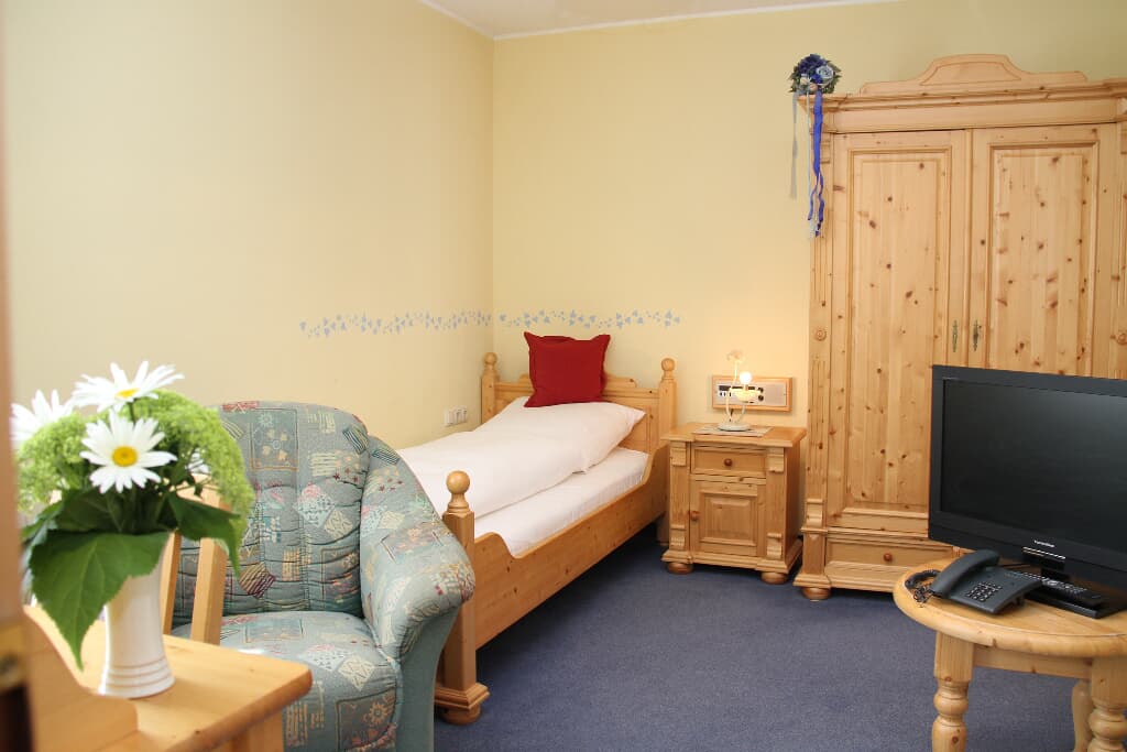 A guest room features light yellow walls and a cozy single bed with white bedding. A wooden wardrobe and desk complement the space. A small TV is positioned on the desk, and a patterned armchair adds a touch of comfort. A decorative flower arrangement sits on the table.