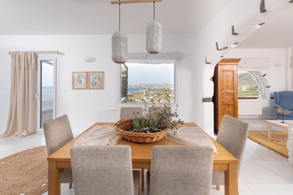A dining area is presented with a wooden table surrounded by four light grey chairs. Above, two textured pendant lights hang. A woven basket filled with greenery sits at the center, while large windows showcase a view of the sea and surrounding landscape.