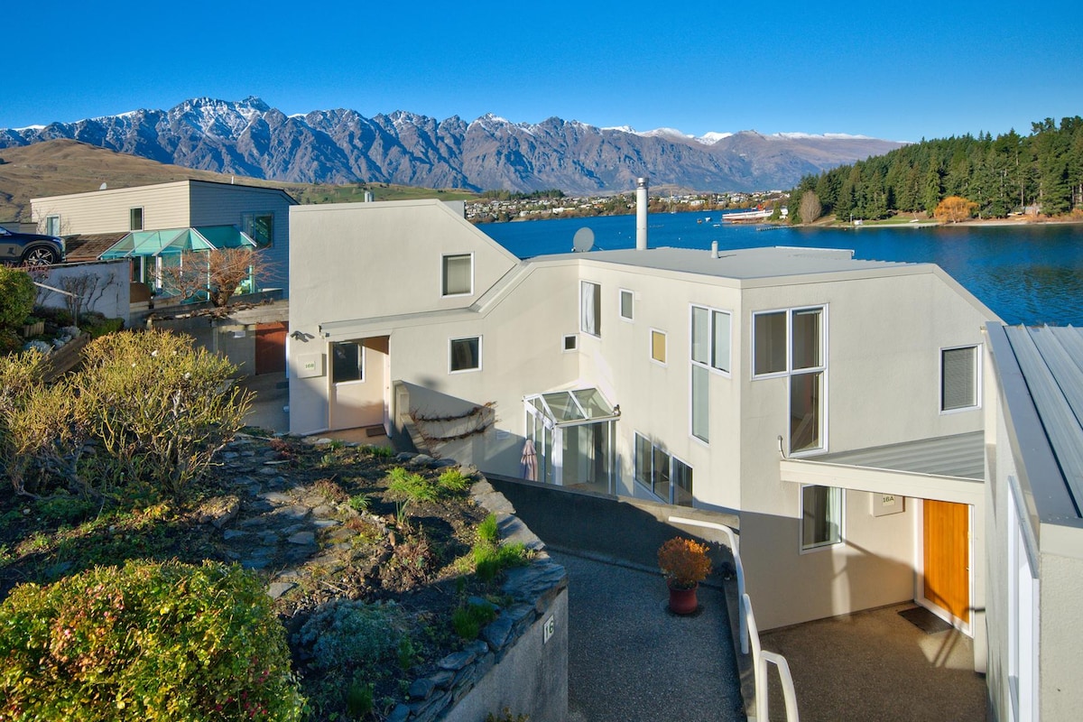 The exterior of the modern home is presented against a backdrop of Lake Wakatipu and the Remarkables mountain range. Large windows reflect the surroundings, while the entrance is framed by a welcoming pathway. Bright sunlight enhances the clean lines and contemporary architecture.