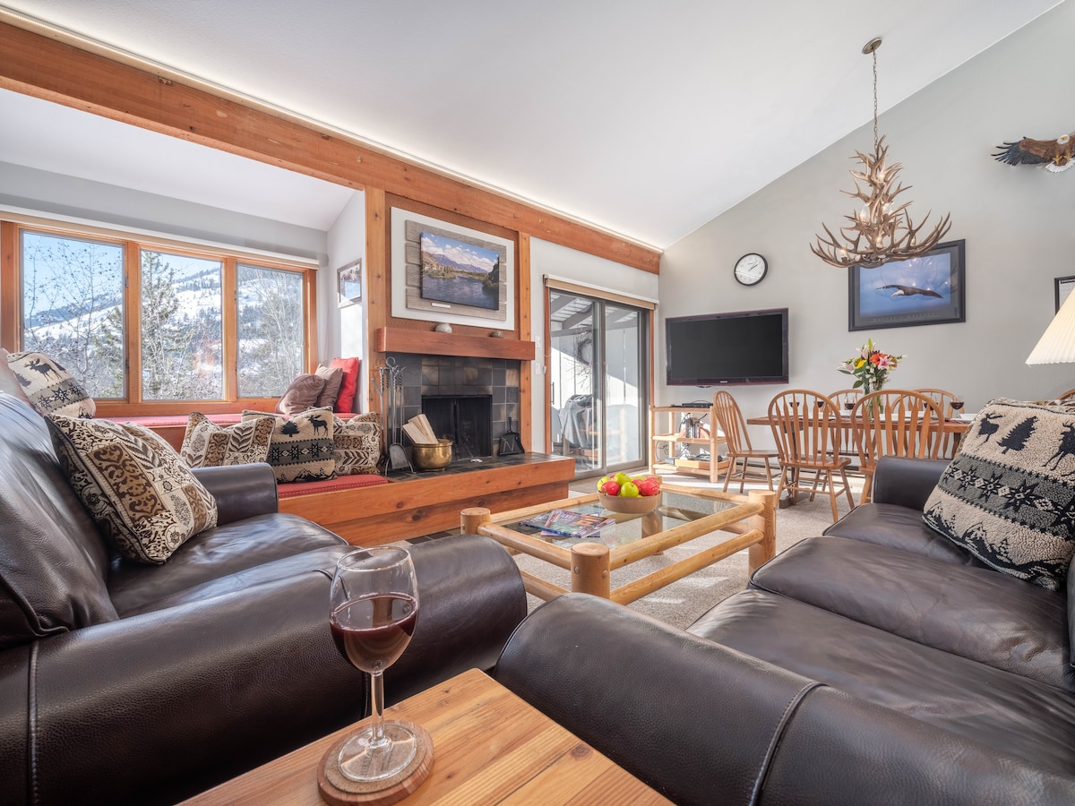 The living room features comfortable leather seating, with a coffee table made of wood. Large windows allow natural light to fill the space, revealing a view of the outdoors. A stone fireplace serves as a focal point, complemented by a dining area visible in the background.