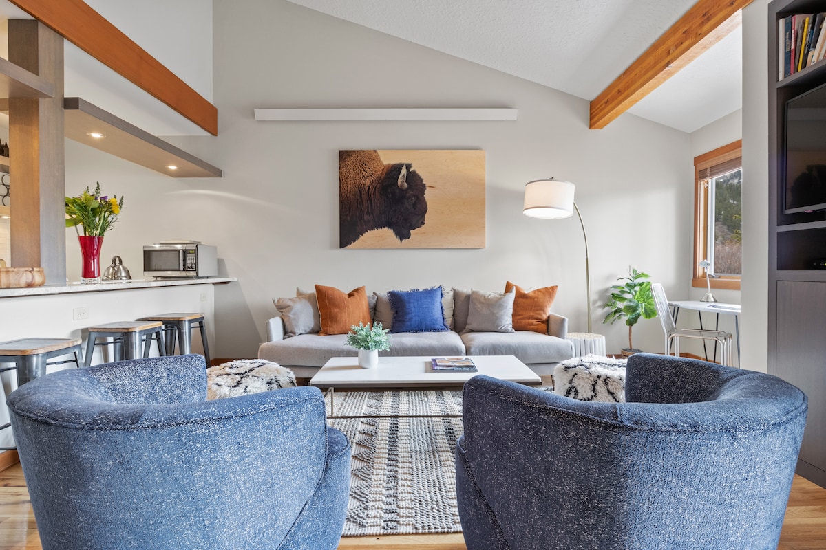 The living area features a modern sofa adorned with colorful pillows, complemented by two blue accent chairs. A coffee table sits on a textured rug, and an artistic buffalo painting hangs above the sofa. Natural light enters through a window, enhancing the bright ambiance.