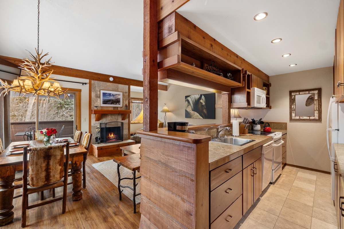 A spacious and well-equipped kitchen features wooden cabinetry and a large countertop. Appliances include a microwave and oven, while a dining area with rustic wooden chairs invites gatherings. Natural light streams in through windows, complemented by soft lighting and a warm wood-burning fireplace in the adjacent living area.