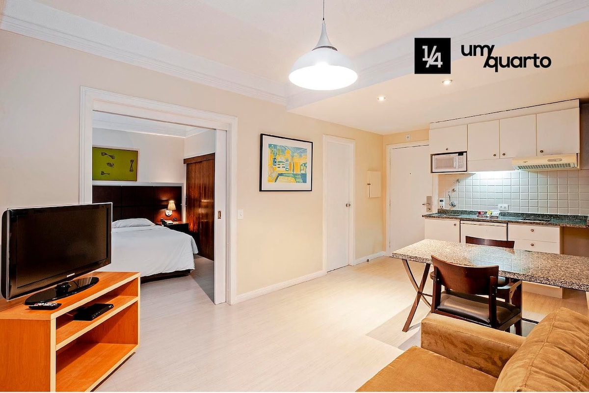 An open layout is displayed, featuring a living area with a television and dining table alongside an American-style kitchenette. A doorway leads to the sleeping area, where a well-made bed is visible. Warm lighting enhances the neutral tones throughout the space.