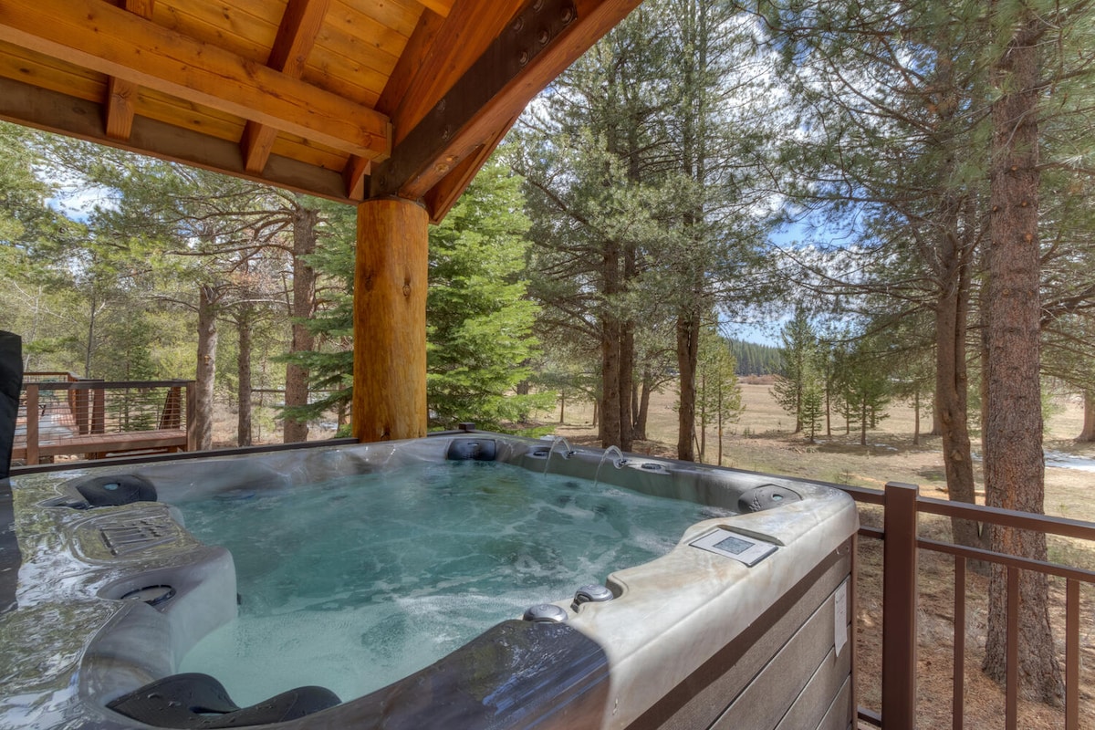 A private hot tub is situated on a wooden deck, surrounded by tall green trees. The bubbling water invites relaxation, while the serene natural landscape stretches into the background, offering a peaceful view of the meadow.