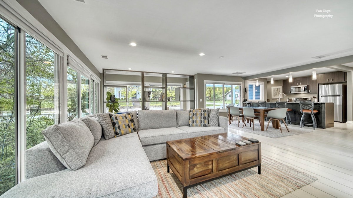 A spacious living area is filled with natural light from surrounding windows. A large sectional sofa, complemented by decorative pillows, creates a comfortable seating arrangement. A wooden coffee table sits in the center, while an open dining space and modern kitchen are visible in the background.