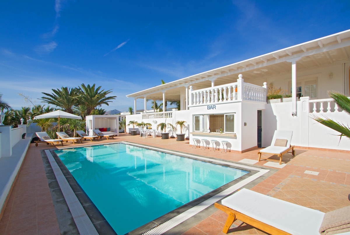 A long heated pool is surrounded by a tiled terrace, with comfortable loungers facing palm trees and clear blue skies. A bar area is visible, complementing the villa's outdoor space. The two-story structure showcases a large balcony with white railings, providing additional seating.