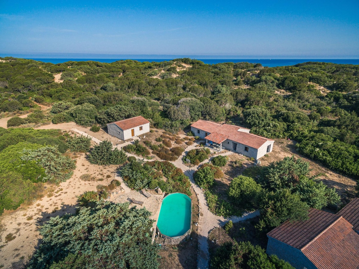 An aerial view captures the two granite buildings with red-tiled roofs, surrounded by lush Mediterranean vegetation. A shared swimming pool nestled in a garden area reflects the blue sky, while the nearby coastline can be seen in the distance.
