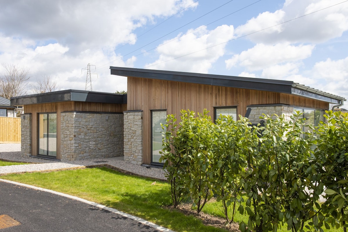 The exterior of the eco retreat is characterized by a blend of natural stone and wooden cladding. Large windows allow natural light to enter. The surrounding area features well-maintained landscaping with greenery and gravel paths, providing a welcoming approach.