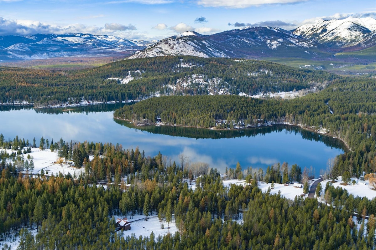 A scenic view of a tranquil lake surrounded by evergreen trees and snow-covered landscapes. Majestic mountains rise in the background, and patches of blue sky are visible, reflecting softly on the water's surface, creating a serene atmosphere.