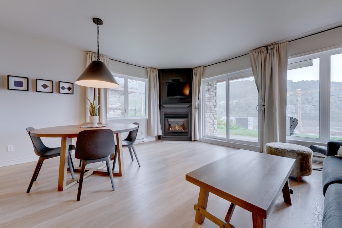 A bright dining area features a round wooden table surrounded by black chairs. Large windows allow natural light to fill the space, while a cozy fireplace is positioned against a stone wall. Soft curtains frame the view of the mountain landscape.