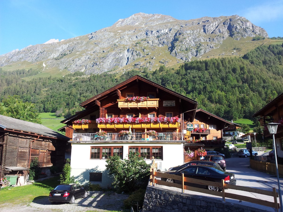 The traditional chalet-style building is surrounded by lush greenery and towering mountains. Balconies adorned with vibrant flower boxes are visible on the upper levels, while a mix of parked vehicles is situated in the foreground, adding to the property's welcoming ambiance.