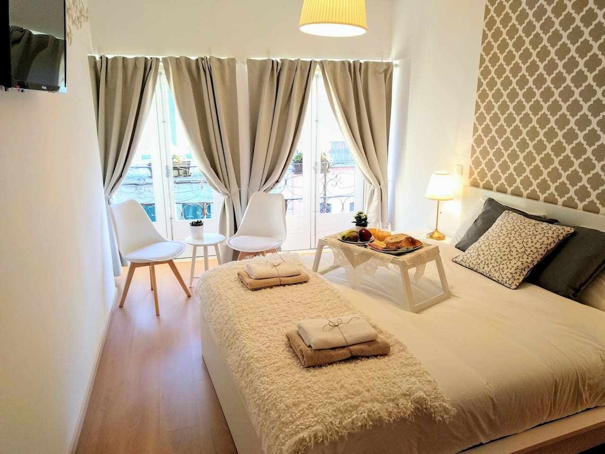 A bright bedroom is illuminated by natural light from two large windows dressed in light curtains. A spacious bed is adorned with soft linens, while a cozy throw blanket adds texture. Two chairs beside a small table create a welcoming seating area.
