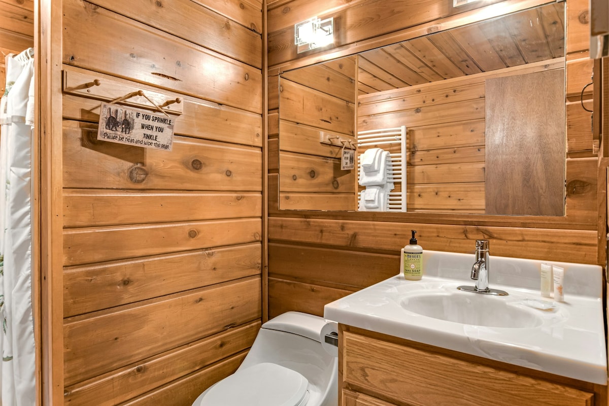 A rustic bathroom features natural wood paneling and a large mirror above a white countertop sink. A flushing toilet is positioned below the mirror, with neatly arranged towels displayed on a rack nearby. Soft lighting enhances the warm atmosphere.