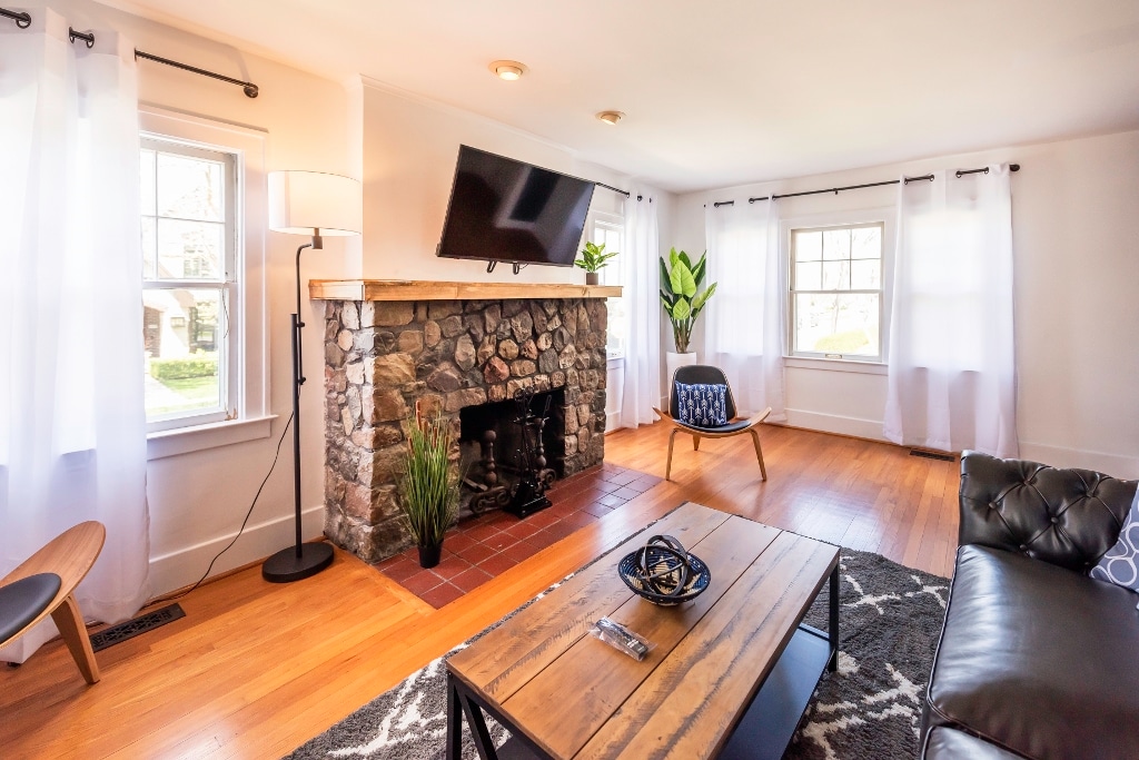 A cozy living area is highlighted by a stone fireplace, which features a mounted television above. Natural light enters through two windows, creating a bright atmosphere. A wooden coffee table stands in the center, accompanied by modern seating and a decorative plant adds life to the space.