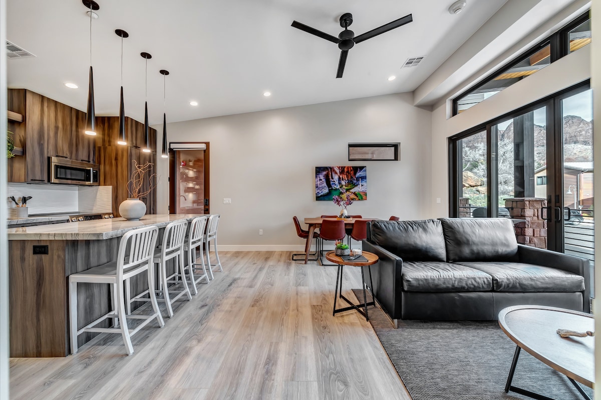 The open-concept living space features a modern kitchen with a large island and bar seating. A comfortable sofa and chairs are positioned near large glass doors that offer views of the surrounding landscape, while stylish pendant lights hang above the kitchen area.