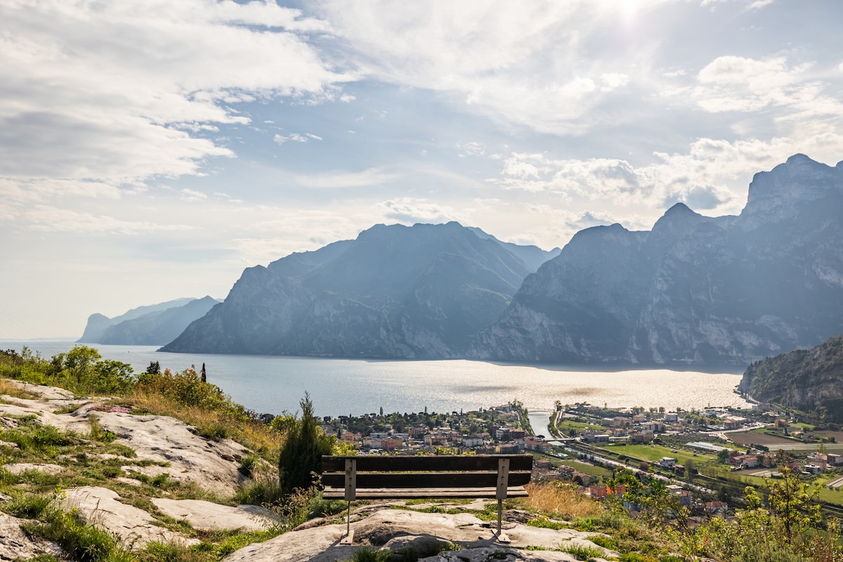 A wooden bench is positioned on a rocky outcrop, overlooking the expansive view of Lake Garda. The scene captures the mountains in the background, with sunlight reflecting on the water. A tranquil atmosphere is created by the natural surroundings and open sky.