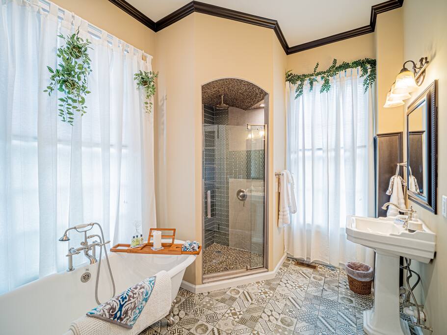 The bathroom features a clawfoot tub complemented by a decorative tray with toiletries. A spacious walk-in shower with modern tiles is visible. Natural light enters through large, sheer-curtained windows, adorned with greenery. Elegant light fixtures illuminate the room, enhancing its charm and functionality.