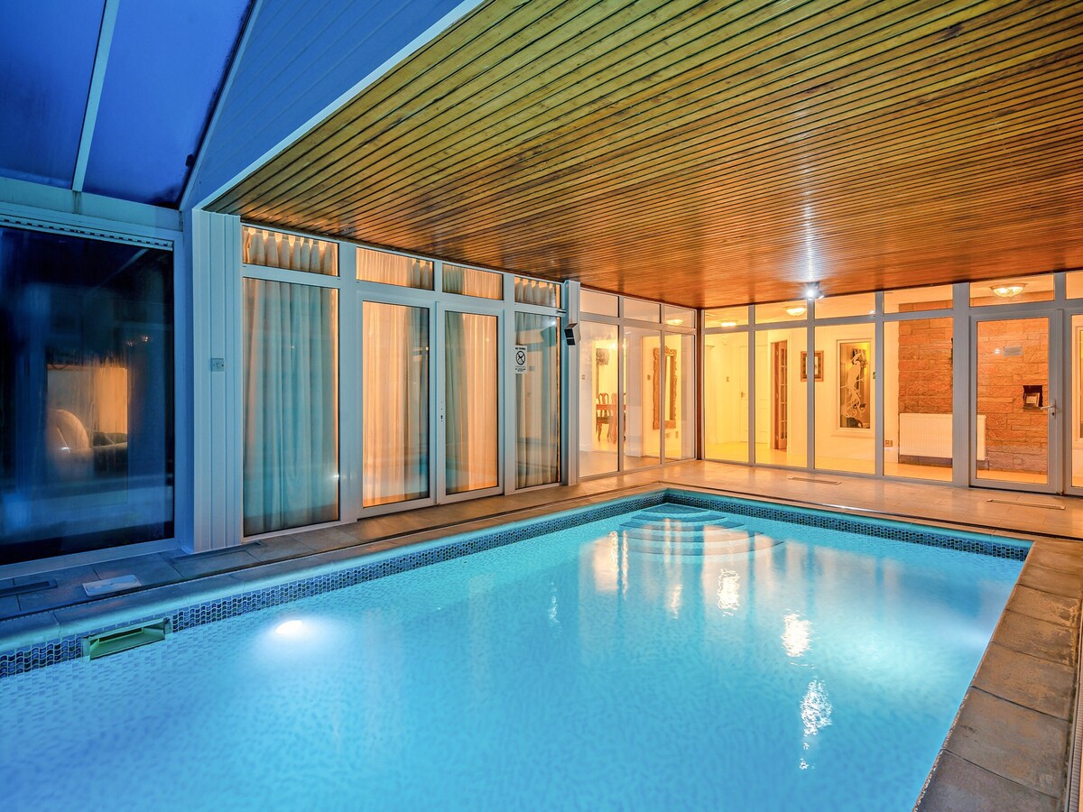 An indoor heated swimming pool is framed by large glass doors that provide a view of the surrounding area. The pool's tranquil water is complemented by a wooden ceiling, creating a spacious and inviting environment. Soft lighting enhances the serene ambiance.