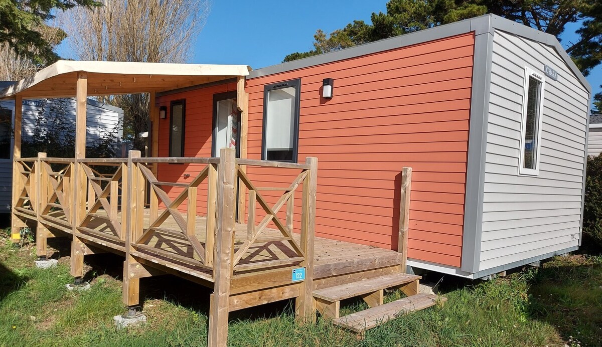 The exterior of the mobile home features a combination of orange and gray siding. A wooden deck with a railing leads up to a semi-covered terrace, providing a welcoming entryway. The structure is surrounded by well-kept grass and trees in the background.