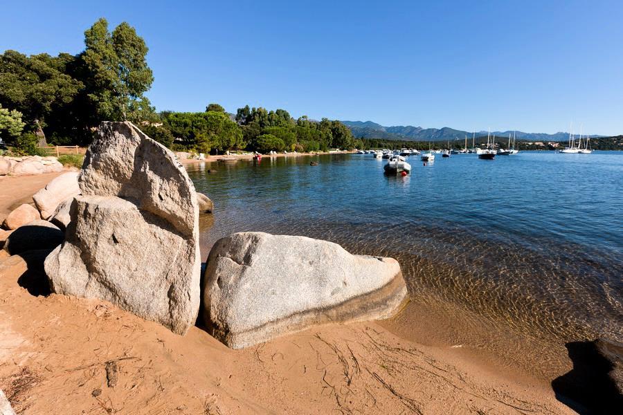 A tranquil sandy beach is framed by large rocks, with calm waters stretching towards the horizon. Several boats are anchored in the bay, surrounded by lush greenery and distant mountains, creating a peaceful coastal setting.