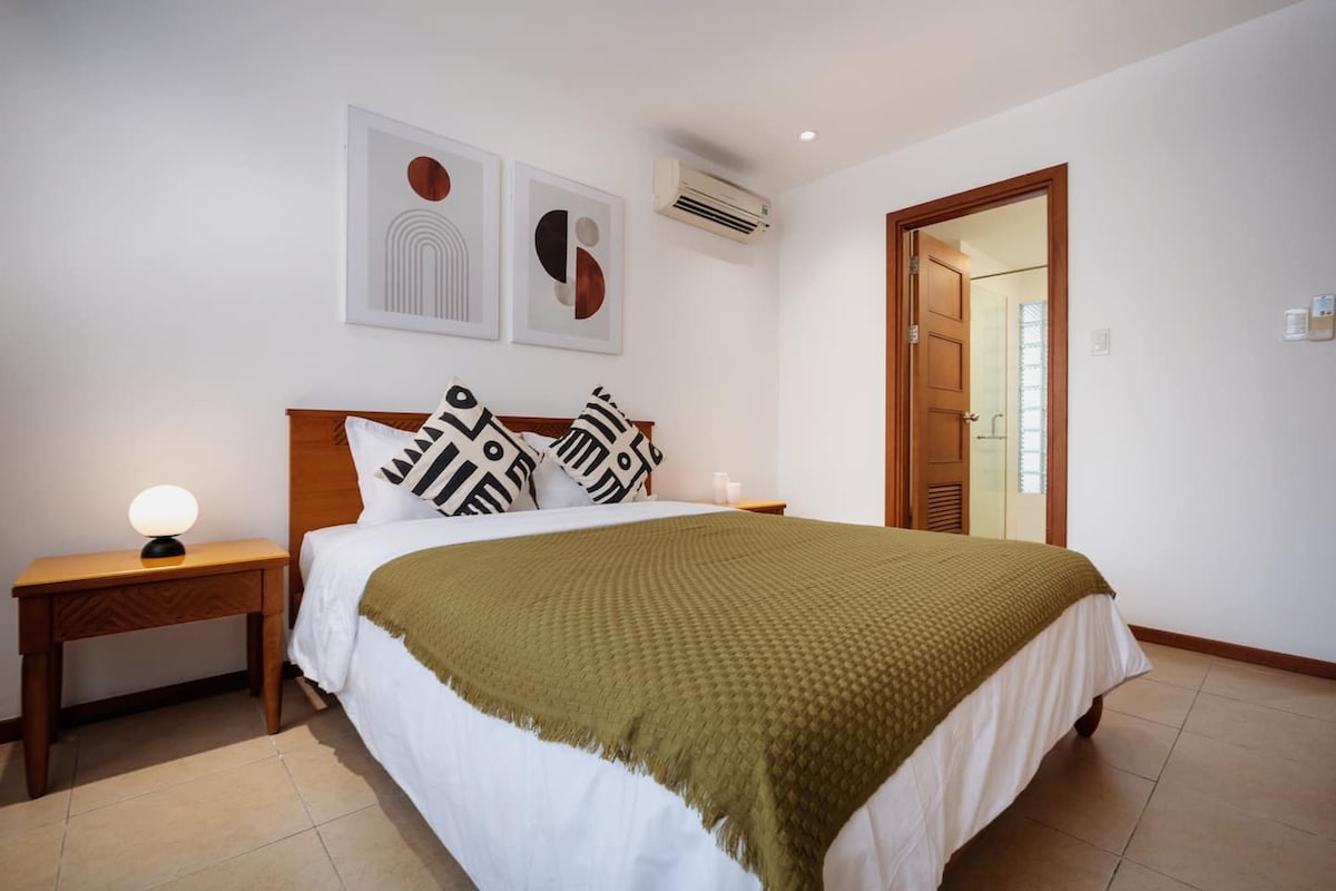 A cozy bedroom is furnished with a spacious bed adorned with a green blanket and decorative pillows. Two modern wall art pieces complement the neutral walls. An air conditioning unit is mounted above the bed, and soft lighting is provided by a bedside lamp on a wooden table.