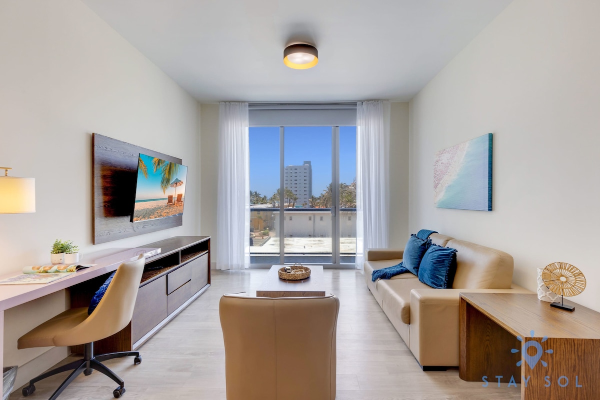 A modern living area is presented with a comfortable pullout sofa and a stylish armchair. A wooden desk is positioned beside a large window, allowing natural light to fill the space. An entertainment unit with a flat-screen TV is mounted on the wall.