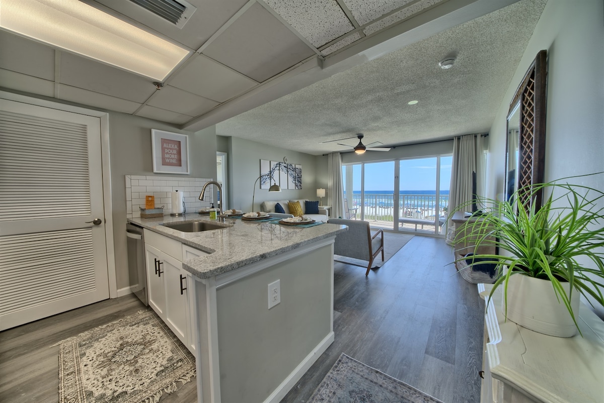 The room features an open floor plan with a kitchen island, complemented by modern cabinetry and a large granite countertop. A ceiling fan adds comfort, while floor-to-ceiling glass doors lead to a private balcony with an ocean view, bringing natural light into the space.